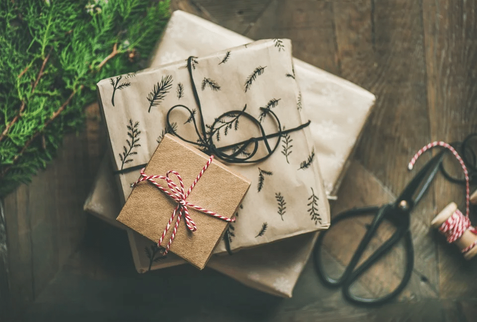 3 grå och bruna presentförpackningar, omgivna av presentband, placerade på ett brunt trägolv med en mejsel och en grankvist i bakgrunden