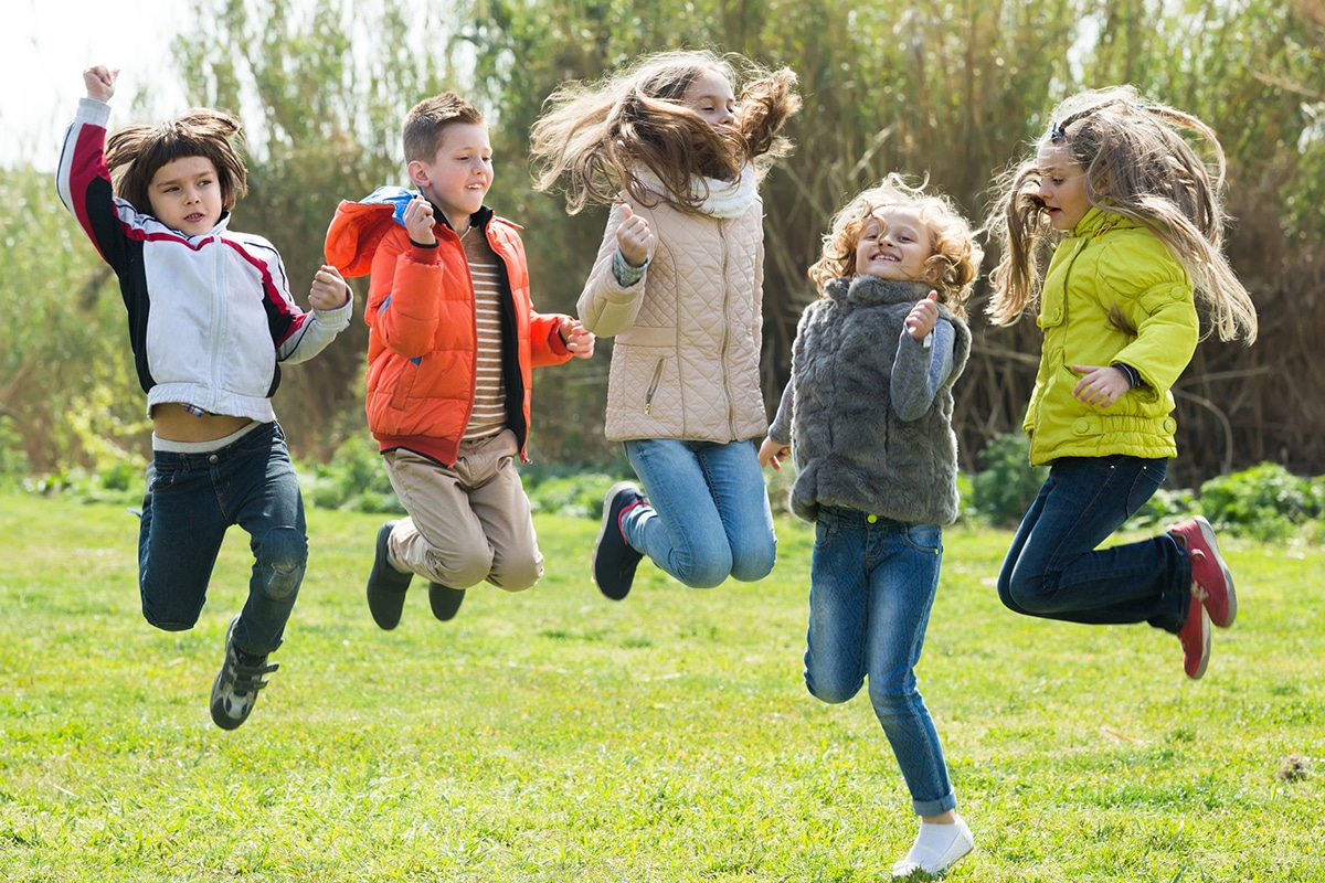 En grupp glada barn som hoppar i gräset utomhus. Det är 2 pojkar och 3 flickor. Det finns träd i bakgrunden