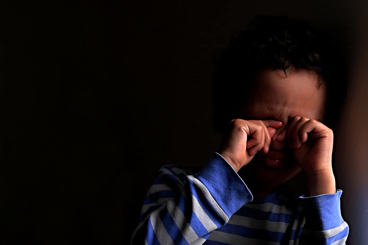 En liten pojke av blandad ras i en blå- och vitrandig tröja. Pojken håller för ögonen eftersom han är rädd för mörkret.
