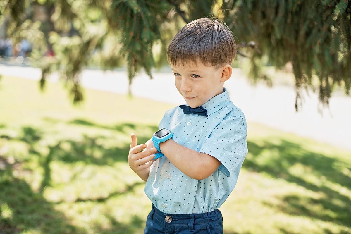 En ung pojke pratar i sin uppkopplade klocka. Han har på sig en blå klocka, en blå skjorta och är ute i solen. Det finns träd i bakgrunden