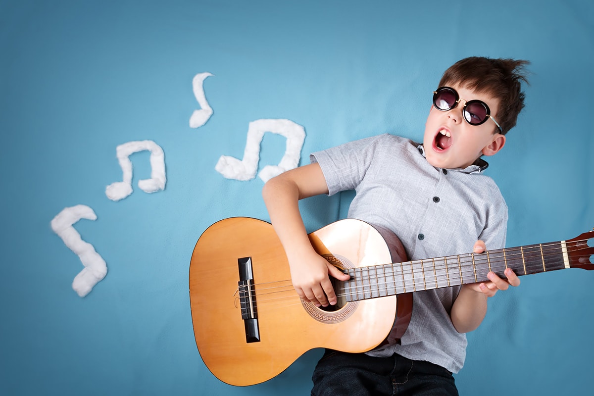 En ung pojke spelar gitarr mot en blå bakgrund. Pojken har mörka glasögon, en grå kortärmad skjorta och en brun trägitarr. Det finns vita noter i bakgrunden