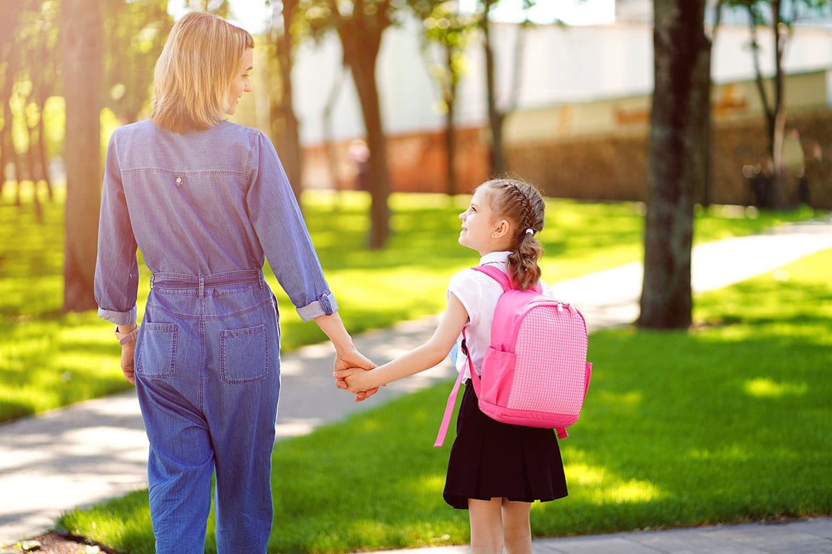 En mamma tar med sin dotter till en park. Det är soligt, mamman är klädd i blå jeans och den lilla flickan har en rosa barnryggsäck