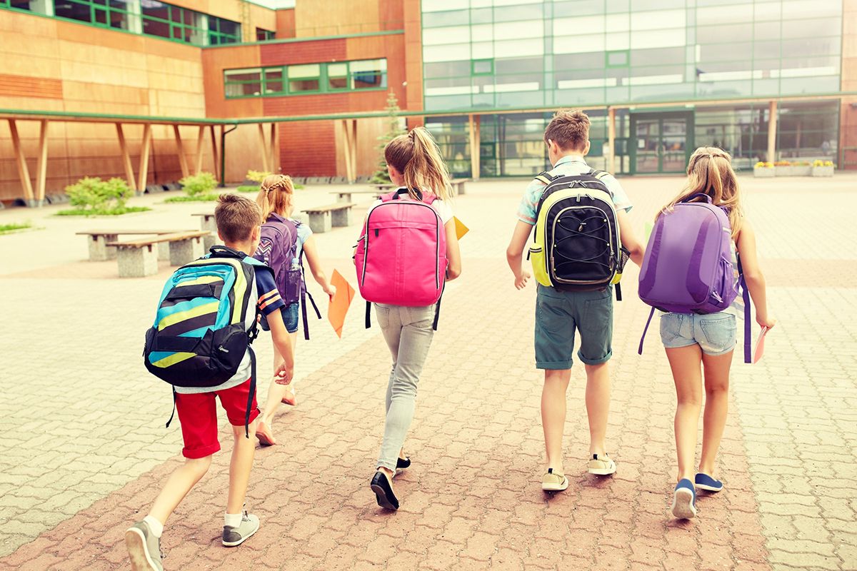 En grupp på 5 barn med ryggen mot skolan. Varje barn bär på en färgad skolväska eller ryggsäck