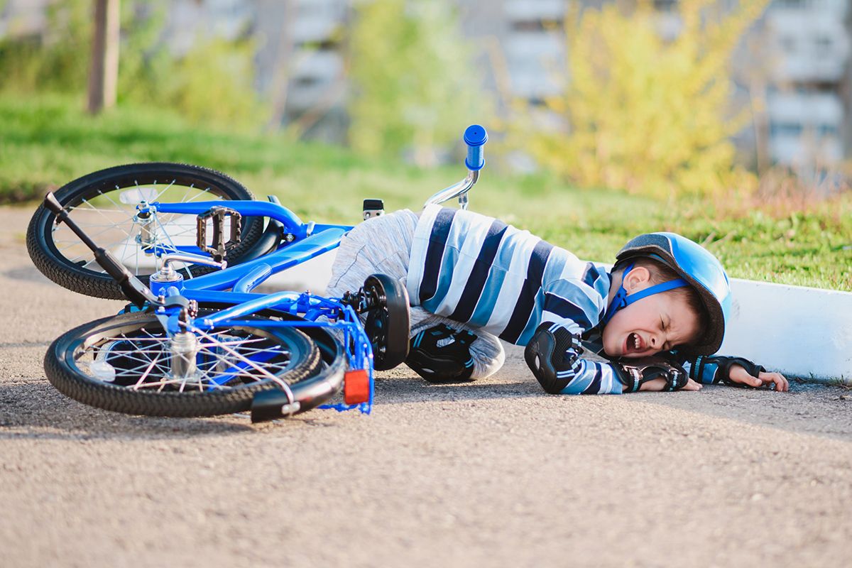 Ett litet barn som har ramlat av sin blå cykel. Han gråter och har på sig cykelhjälm, knäskydd och svarta skyddshandskar
