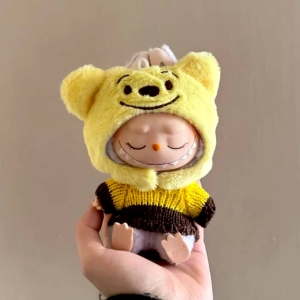 Klädset för gosedjur med gul huva med leende öron och randig gul-brun tröja, buren av en sovande Labubu-docka.